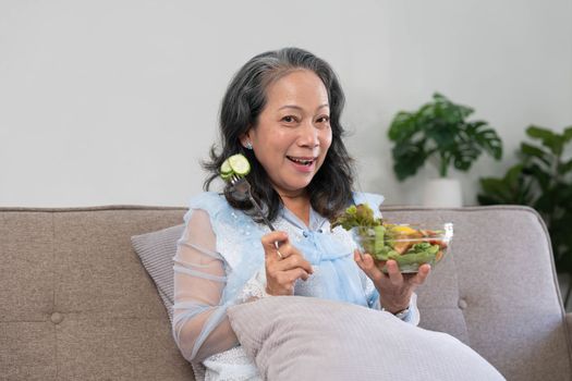 Portrait, Cheerful Asian 60s aged woman having breakfast in her living room, eating healthy salad vegetables mix bowl...