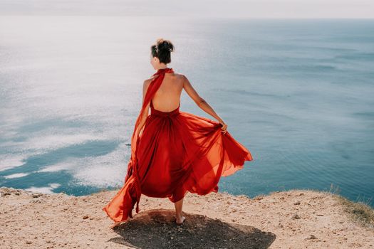 Side view a Young beautiful sensual woman in a red long dress posing on a rock high above the sea during sunrise. Girl on the nature on blue sky background. Fashion photo.