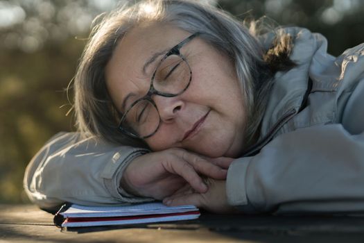 older white-haired woman with glasses asleep on her notebook in the park dreaming