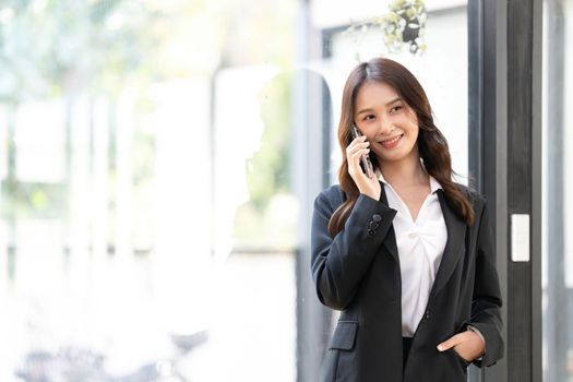 Happy young asian business woman wearing suit holding mobile phone standing in her workstation office..