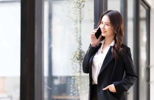 Happy young asian business woman wearing suit holding mobile phone standing in her workstation office..