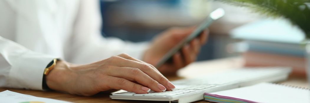 Female hands hold smartphone and work on laptop keyboard. Mobile applications for business and analytics