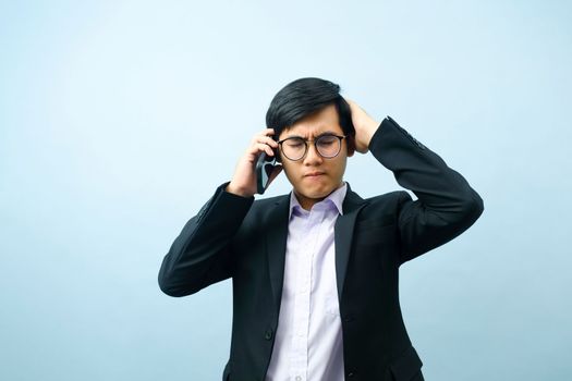 Portrait of young smart asian businessman in eyeglasses standing and talking on mobile phone stressed and frustrated about business with light blue isolated background. Business and connection concept.