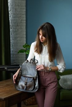 beautiful slender girl in burgundy pants and a white blouse posing with a brown leather backpack. Indoor photo