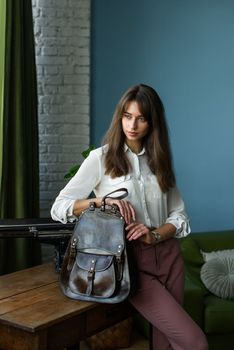 beautiful slender girl in burgundy pants and a white blouse posing with a brown leather backpack. Indoor photo