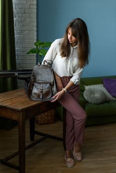 beautiful slender girl in burgundy pants and a white blouse posing with a brown leather backpack. Indoor photo