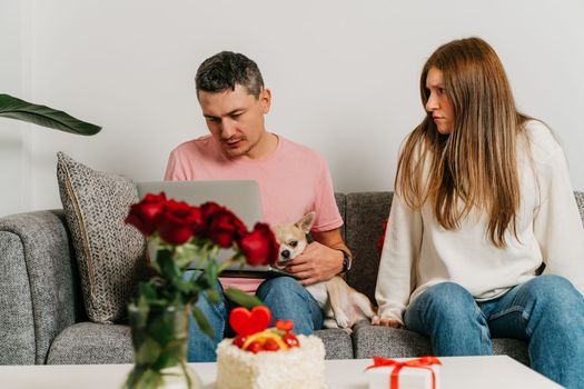 Beautiful young couple with dog sitting on a couch, having a quarrel over romantic date on Valentine day. Man working at laptop, woman in anger.