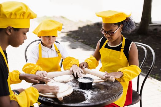 Multiethnic cooks children in yellow chefs hat and apron cooking dough for bakery. Black african and caucasian child cooking and having fun together