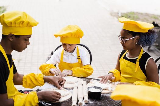 Multiethnic cooks children in yellow chefs hat and apron cooking dough for bakery. Black african and caucasian child cooking and having fun together