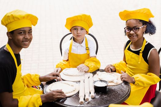 Multiethnic cooks children in yellow chefs hat and apron cooking dough for bakery. Black african and caucasian child cooking and having fun together