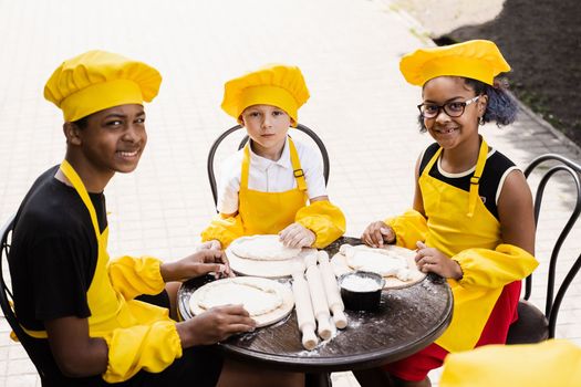 Multiethnic cooks children in yellow chefs hat and apron cooking dough for bakery. Black african and caucasian child cooking and having fun together