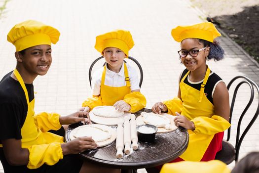 Multiethnic cooks children in yellow chefs hat and apron cooking dough for bakery. Black african and caucasian child cooking and having fun together