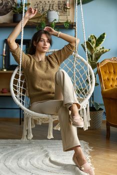 Young attractive woman chilling at home in comfortable hanging chair. beautiful slender girl in beige pants and olive sweater posing while sitting in a swing chair