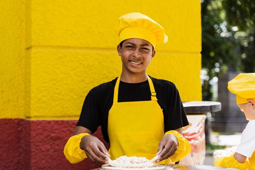 Black african teenager cook in chefs hat and yellow apron uniform cooking dough for bakery. Creative advertising for cafe or restaurant