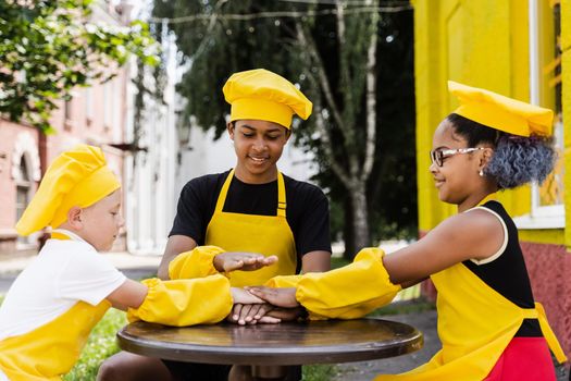 Teambuilding of multinational children cooks in chefs hat and yellow apron uniform put hands on each other, having fun and laughing. Multiethnic kids commutication activity