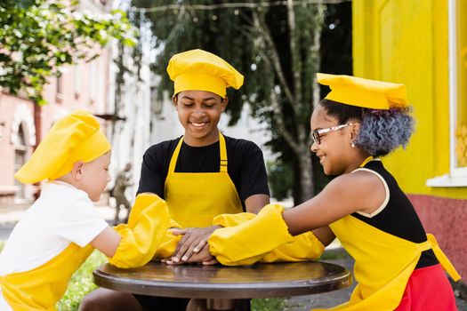 Teambuilding of multinational children cooks in chefs hat and yellow apron uniform put hands on each other, having fun and laughing. Multiethnic kids commutication activity