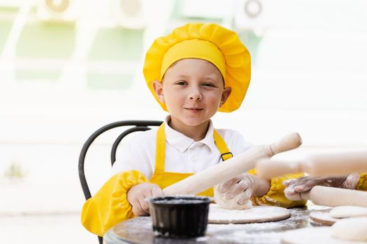 Handsome cook child in yellow chefs hat and apron yellow uniform holding dough roller and cooking dough outdoor