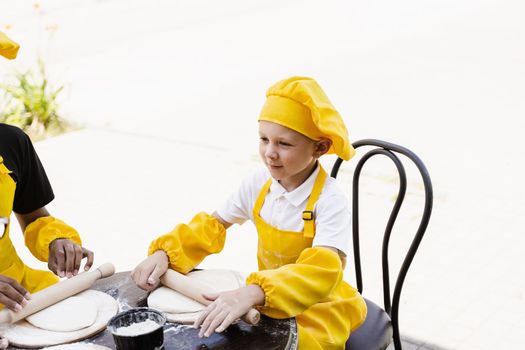 Handsome cook child in yellow chefs hat and apron yellow uniform rolling dough and cooking outdoor