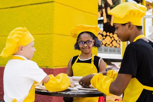 Multinational company of children cooks in yellow uniforms cooking dough for bakery. African teenager and black girl have fun with caucasian child boy and cook food