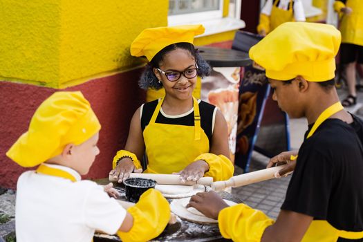 Multinational company of children cooks in yellow uniforms cooking dough for bakery. African teenager and black girl have fun with caucasian child boy and cook food