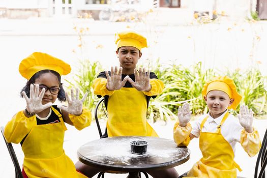 Multiracial children cooks play with flour for dough and having fun. Multinational cook kids in chefs hat and yellow apron uniform cooking outdoor for bakery