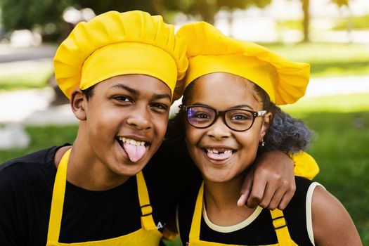 African children cooks in chefs hat and yellow uniforms grimacing and showing tongue each others. African teenager and black girl have fun and cook food