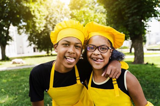 African children cooks in chefs hat and yellow uniforms grimacing and showing tongue each others. African teenager and black girl have fun and cook food