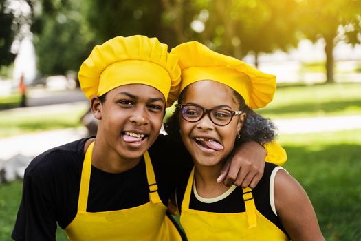 African children cooks in chefs hat and yellow uniforms grimacing and showing tongue each others. African teenager and black girl have fun and cook food