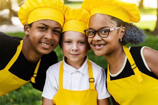Multiracial company of children cooks in yellow uniforms smile outdoor. African teenager and black girl have fun with caucasian child boy and cook food. Kids portrait