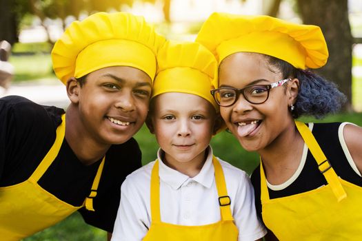 Multinational company of children cooks in yellow uniforms smiling and grimacing and showing tongue outdoor. African teenager and black girl have fun with caucasian child boy and cook food