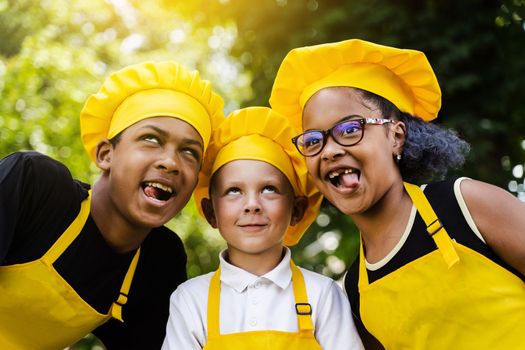 Multinational company of children cooks in yellow uniforms smiling and grimacing and showing tongue outdoor. African teenager and black girl have fun with caucasian child boy and cook food