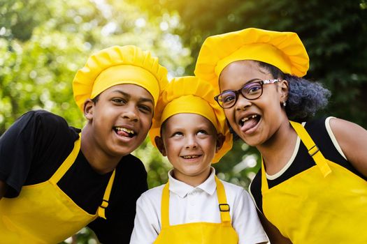 Multinational company of children cooks in yellow uniforms smiling and grimacing and showing tongue outdoor. African teenager and black girl have fun with caucasian child boy and cook food