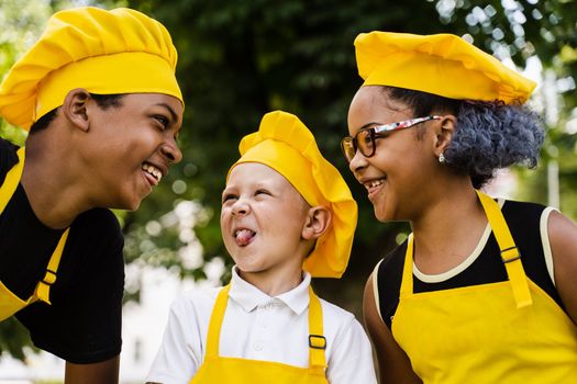 Multinational company of children cooks in yellow uniforms smiling and grimacing and showing tongue outdoor. African teenager and black girl have fun with caucasian child boy and cook food