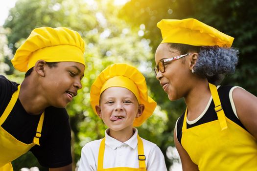 Multinational company of children cooks in yellow uniforms smiling and grimacing and showing tongue outdoor. African teenager and black girl have fun with caucasian child boy and cook food