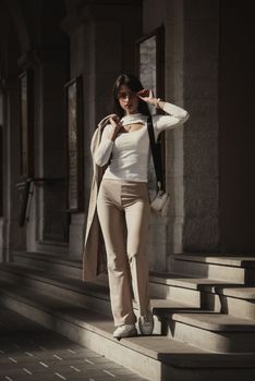 beautiful brunette girl dressed in white blouse, pants, sneakers with a handbag and camel coat in hands. Stylish trendy fashion outlook, full length lifestyle model