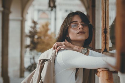 beautiful brunette girl in a sunglasses dressed in white blouse with a beige coat in hands. Stylish trendy fashion outlook.