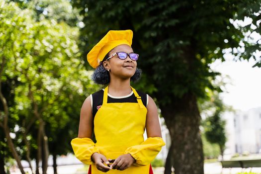 Black african child cook girl in chefs hat and yellow apron uniform smiling outdoor. Creative advertising for cafe or restaurant