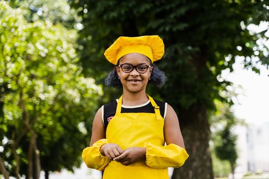 Black african child cook girl in chefs hat and yellow apron uniform smiling outdoor. Creative advertising for cafe or restaurant