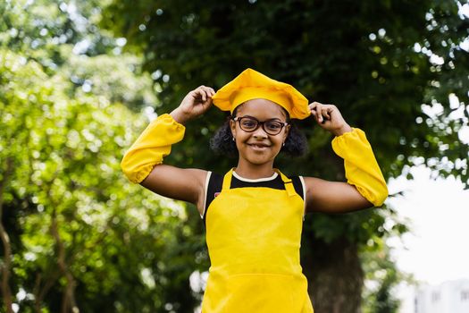 Black african child cook girl in chefs hat and yellow apron uniform touching her chefs hat. Creative advertising for cafe or restaurant