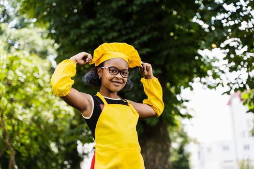 Black african child cook girl in chefs hat and yellow apron uniform touching her chefs hat. Creative advertising for cafe or restaurant
