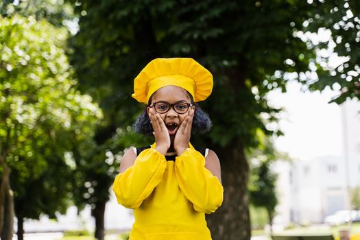 Shocked black african child cook girl in chefs hat and yellow apron uniform hold cheeks and surprise. Creative advertising for cafe or restaurant