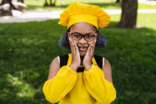 Shocked black african child cook girl in chefs hat and yellow apron uniform hold cheeks and surprise. Creative advertising for cafe or restaurant