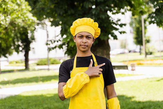 Handsome african teenager cook points right side. Black child cook in chefs hat and yellow apron uniform smiling and pointing right side outdoor. Creative advertising for cafe or restaurant