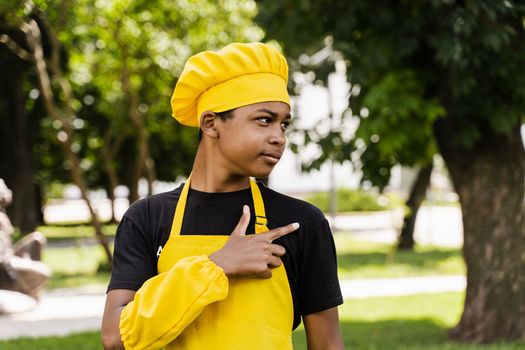 Handsome african teenager cook points right side. Black child cook in chefs hat and yellow apron uniform smiling and pointing right side outdoor. Creative advertising for cafe or restaurant