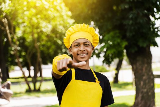 Handsome african teenager cook points to you. Black child cook in chefs hat and yellow apron uniform smiling and pointing to you outdoor. Creative advertising for cafe or restaurant.