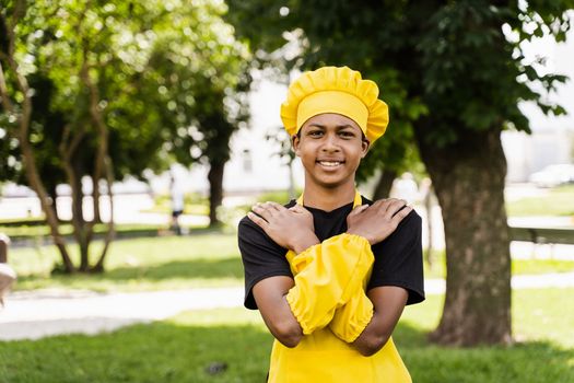 Black african teenager cook in chefs hat and yellow apron uniform smiling outdoor. Creative advertising for cafe or restaurant