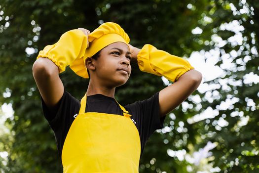 Black african teenager cook in chefs hat and yellow apron uniform touching his chefs hat. Creative advertising for cafe or restaurant