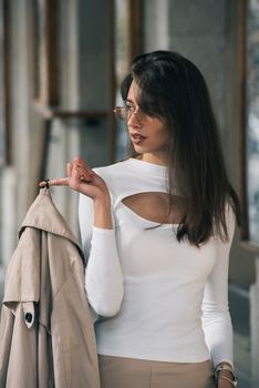 beautiful brunette girl in a sunglasses dressed in white blouse with a beige coat in hands. Stylish trendy fashion outlook.