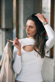 beautiful brunette girl in a sunglasses dressed in white blouse with a beige coat in hands. Stylish trendy fashion outlook.