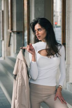 beautiful brunette girl in a sunglasses dressed in white blouse with a beige coat in hands. Stylish trendy fashion outlook.